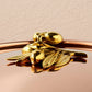 A close-up of a shiny brass decorative olive-branch knob on a reflective copper lid against a light beige background, from the Olivia Cupra Ruffoni collection