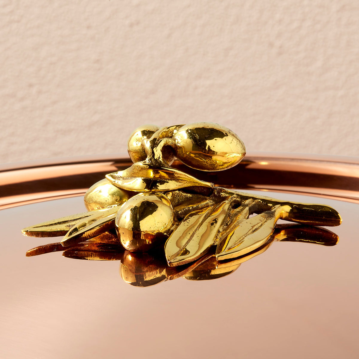 A close-up of a shiny brass decorative olive-branch knob on a reflective copper lid against a light beige background, from the Olivia Cupra Ruffoni collection