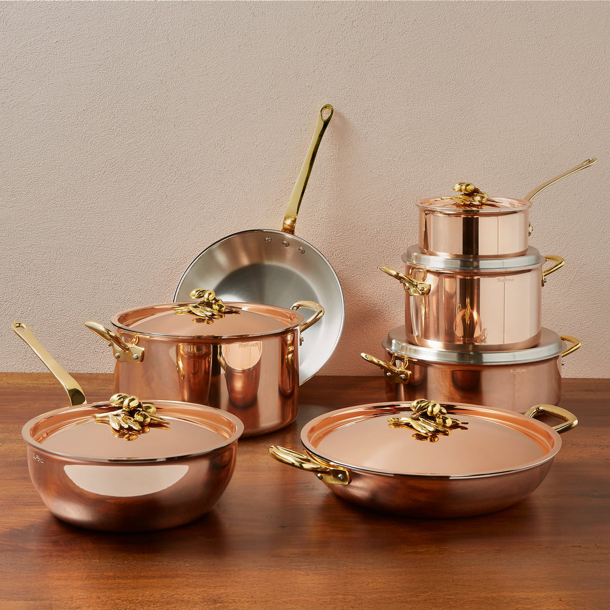 Ruffoni Olivia Cupra copper cookware set with shiny brass handles and lids, arranged on a wooden table against a textured beige wall. The set includes multiple pots, pans, and stacked saucepans.