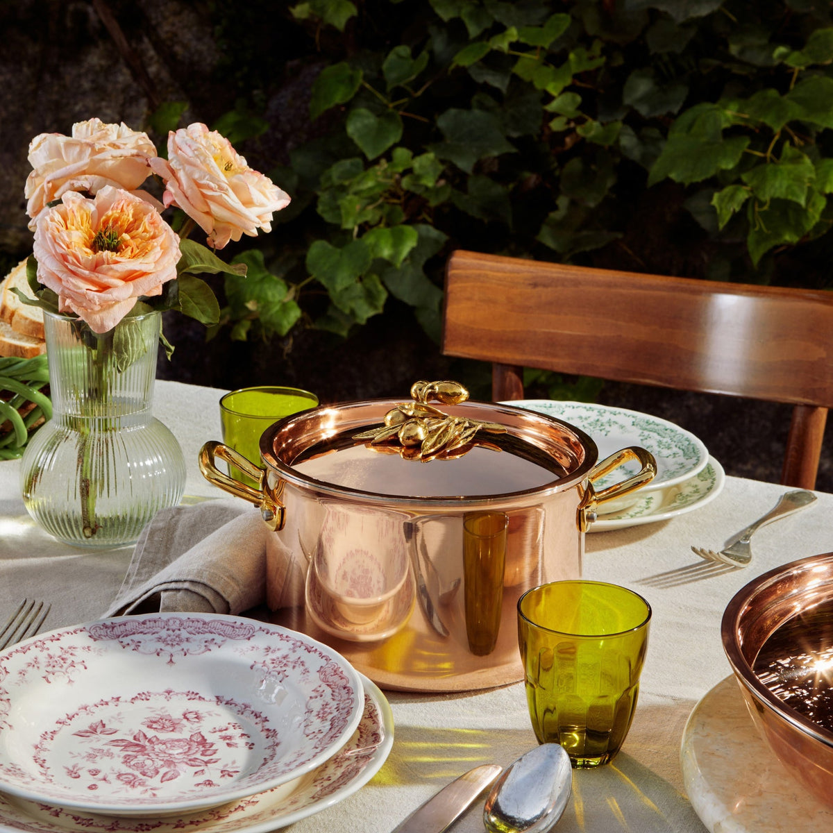 A polished copper soup pot with brass handles and olive branch knob sits on a dining table set with patterned plates, green glass cups, a vase of peach roses, and a wooden chair against a leafy outdoor background