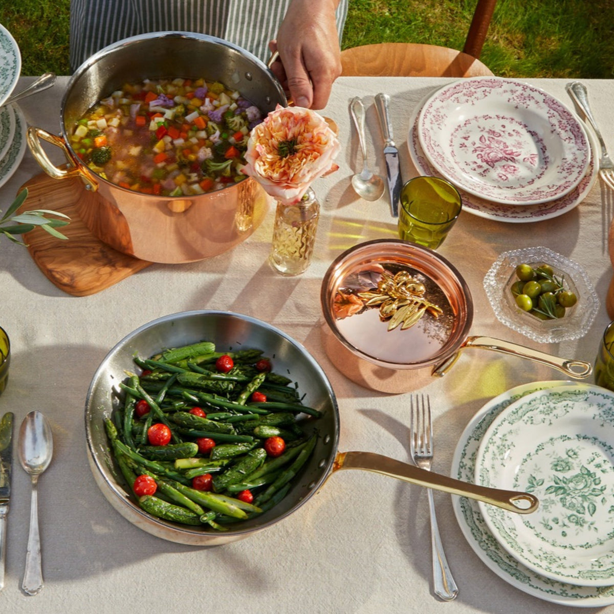 An outdoor dining table styled with Olivia Cupra copper cookware, floral ceramic plates, silver cutlery, and fresh food including asparagus, tomatoes, olives, and a vegetable stew. 