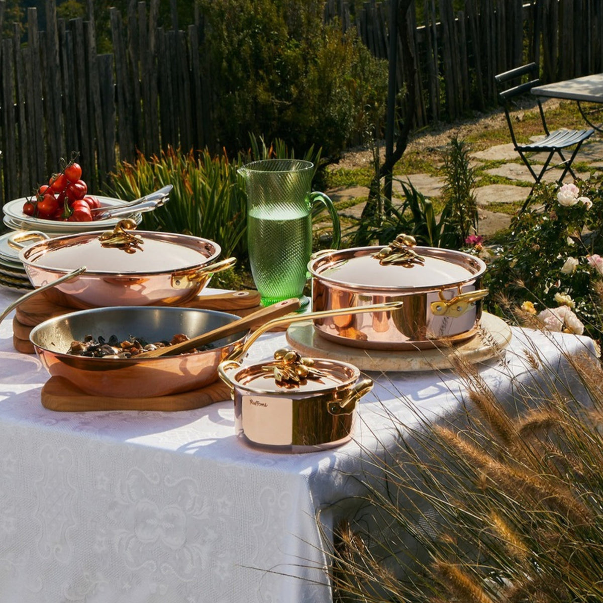 A set of copper cookware with brass handles and lids, arranged on a white tablecloth outdoors in a garden setting. The cookware includes multiple saucepans, a frying pan, and a glass pitcher.