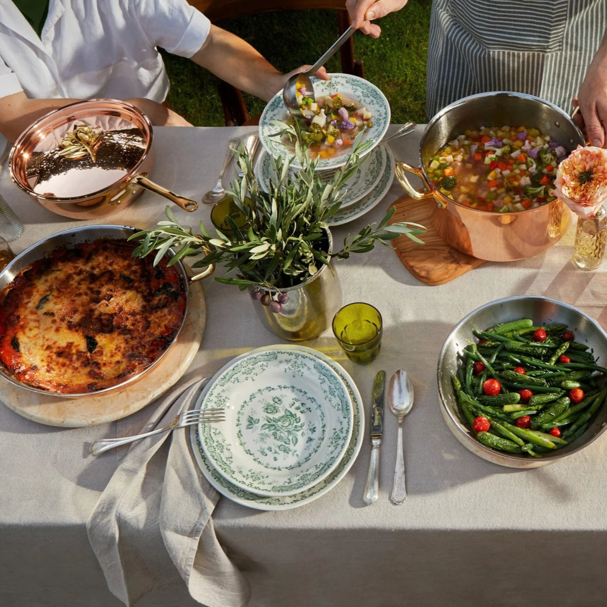 Olivia Cupra 7 piece set on a beige linen tablecloth with green plates and glasses in the Italian countryside on a summer day