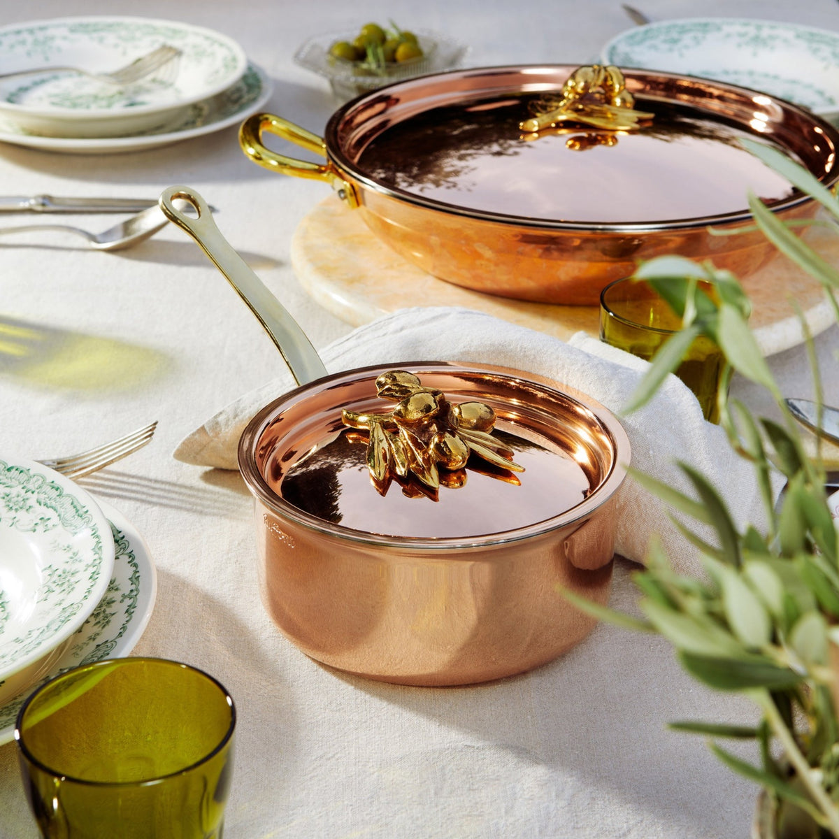 Two Olivia Cupra copper pots with brass handles, styled on a white tablecloth with green glassware, plates, and plants in a bright dining setting